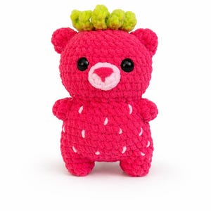 Peut inclure: Un ours fraise en peluche fait à la main. L'ours est rose vif avec des détails de graines blanches et une couronne de feuilles vertes. Il a des yeux noirs et un nez rose. L'ours est debout sur un fond blanc.