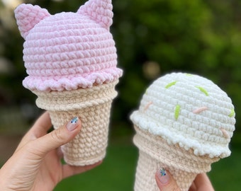 May include: Two crocheted ice cream cones, one pink with cat ears and one white with green and pink sprinkles.