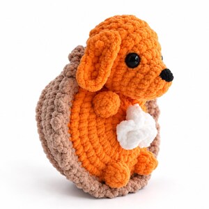 May include: A crocheted plush toy shaped like a dog sitting inside a brown pie crust. The dog is bright orange with black plastic eyes and a black nose. A small white dollop of whipped cream is attached to the dog's chest area.