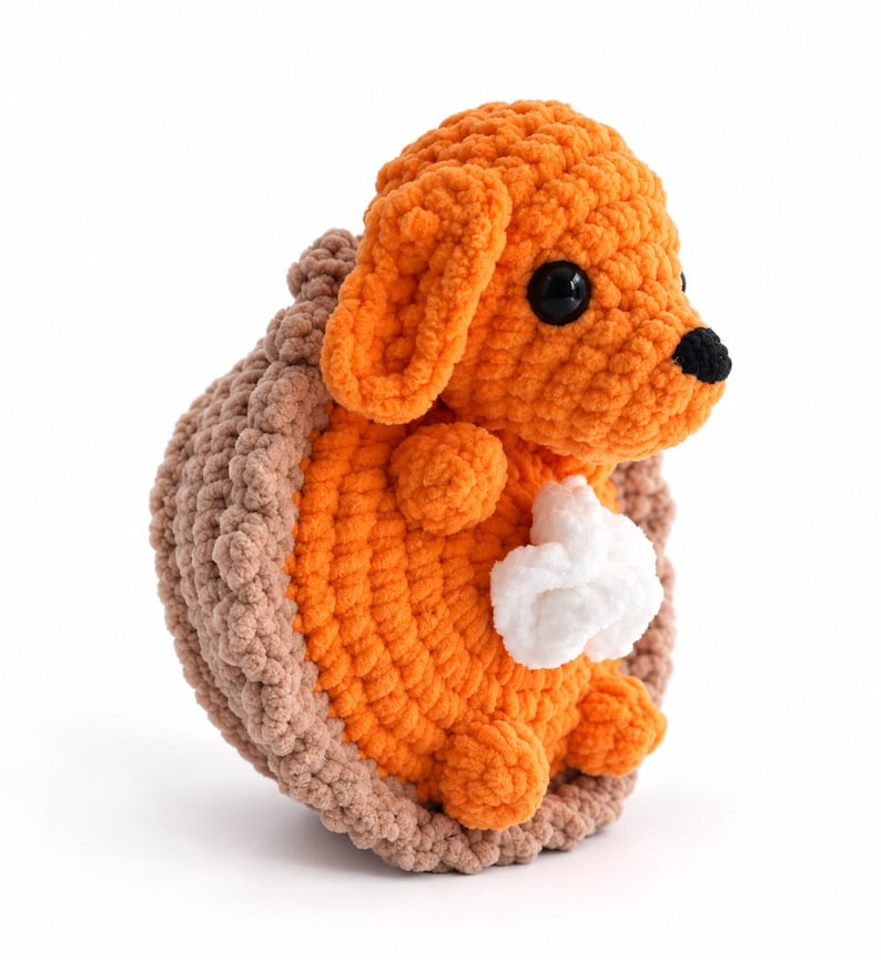 May include: A crocheted plush toy shaped like a dog sitting inside a brown pie crust. The dog is bright orange with black plastic eyes and a black nose. A small white dollop of whipped cream is attached to the dog's chest area.