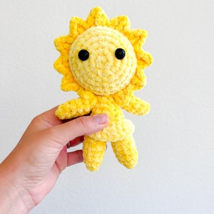 May include: A yellow crocheted sun plush toy with black eyes. The sun has a smiling face and is holding its arms out.