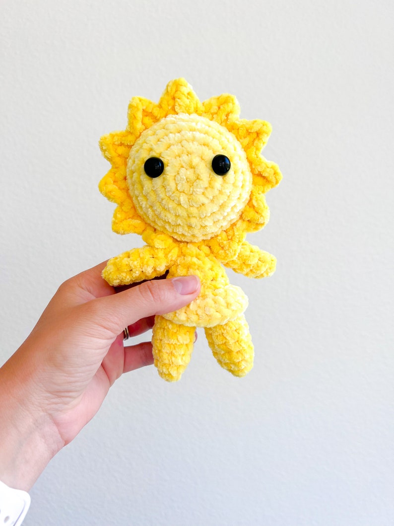 May include: A yellow crocheted sun plush toy with black eyes. The sun has a smiling face and is holding its arms out.