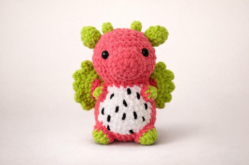 Puede incluir: Un peluche de drag&oacute;n hecho a mano de ganchillo en rosa, verde y blanco. El drag&oacute;n tiene un cuerpo rosa, alas y detalles verdes, y una barriga blanca con detalles negros en forma de semillas. El drag&oacute;n tiene ojos negros.