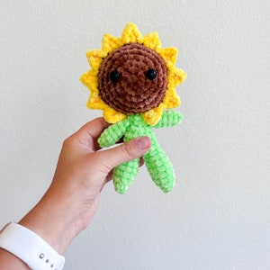 May include: A crocheted sunflower plush toy with a brown face, black eyes, and green limbs. The sunflower has yellow petals.