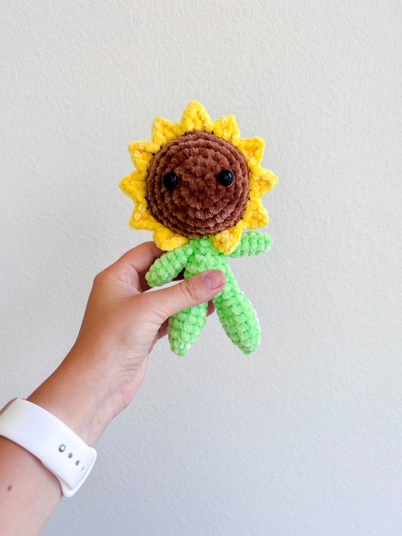 May include: A crocheted sunflower plush toy with a brown face, black eyes, and green limbs. The sunflower has yellow petals.