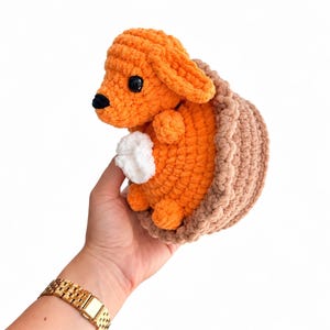 May include: A small, handmade crochet plush toy featuring a bright orange animal with floppy ears, a black nose, and black eyes, nestled inside a textured, light brown crocheted basket-like structure with a small white tuft of yarn on its chest.