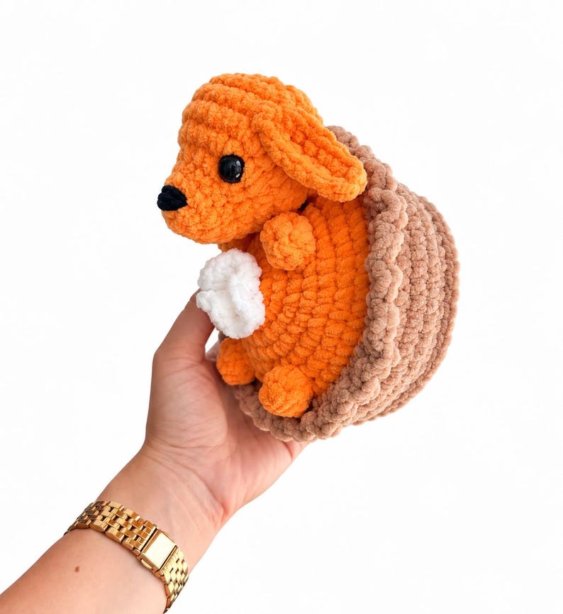 May include: A small, handmade crochet plush toy featuring a bright orange animal with floppy ears, a black nose, and black eyes, nestled inside a textured, light brown crocheted basket-like structure with a small white tuft of yarn on its chest.