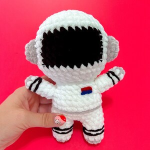 May include: White crocheted astronaut toy with black eyes and a blue and red patch on the chest. The toy is standing on a pink surface.