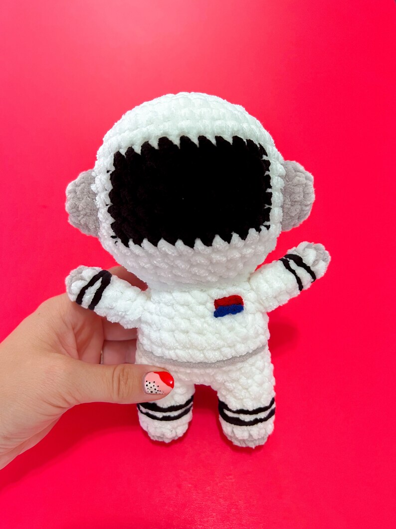 May include: White crocheted astronaut toy with black eyes and a blue and red patch on the chest. The toy is standing on a pink surface.