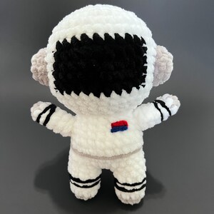 May include: White crocheted astronaut toy with black eyes and a red and blue patch on the chest.