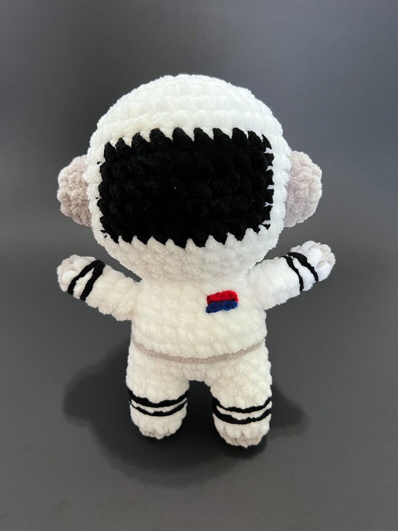 May include: White crocheted astronaut toy with black eyes and a red and blue patch on the chest.