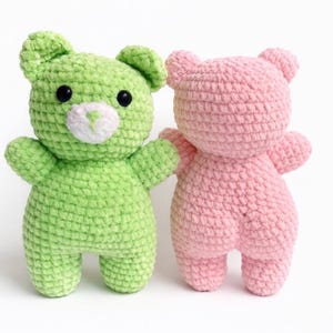 May include: Two handmade crochet teddy bears. One is lime green with black eyes and a white muzzle. The other is light pink. Both have small arms and legs, and are standing upright on a white surface.