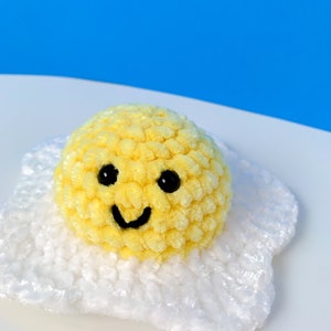 K&ouml;nnte beinhalten: Ein gelbes, geh&auml;keltes Smiley-Ei mit schwarzen Augen und einem L&auml;cheln, das auf einem wei&szlig;en, geh&auml;kelten Eiwei&szlig; sitzt.