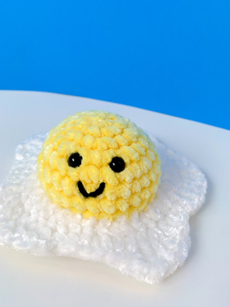 K&ouml;nnte beinhalten: Ein gelbes, geh&auml;keltes Smiley-Ei mit schwarzen Augen und einem L&auml;cheln, das auf einem wei&szlig;en, geh&auml;kelten Eiwei&szlig; sitzt.