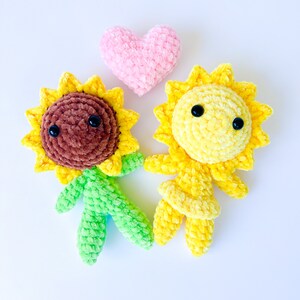 May include: Two crocheted sunflower plushies, one brown and one yellow, with black eyes and a pink heart between them.