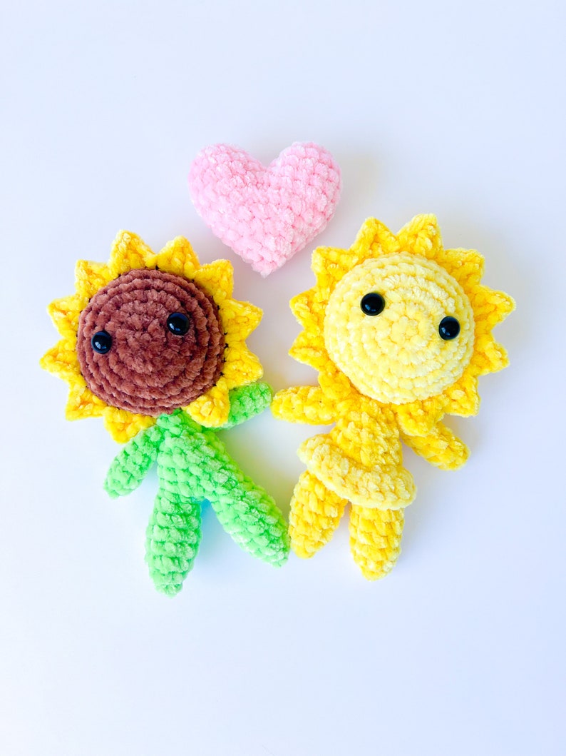 May include: Two crocheted sunflower plushies, one brown and one yellow, with black eyes and a pink heart between them.
