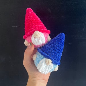 May include: Two crocheted gnomes, one pink and one blue, with white yarn beards. The gnomes are held in a hand against a black background.