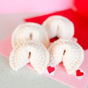 May include: A close-up of four cream-colored, crocheted fortune cookies with a small red heart on a white tag attached to each. The cookies rest on a pink felt surface, with a red background.