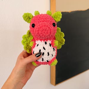 Puede incluir: Un peluche de drag&oacute;n hecho a mano con ganchillo en forma de fruta del drag&oacute;n. El peluche presenta un cuerpo rosa, alas y detalles verdes, y una barriga blanca con detalles de semillas negras. El drag&oacute;n tiene ojos de seguridad negros.