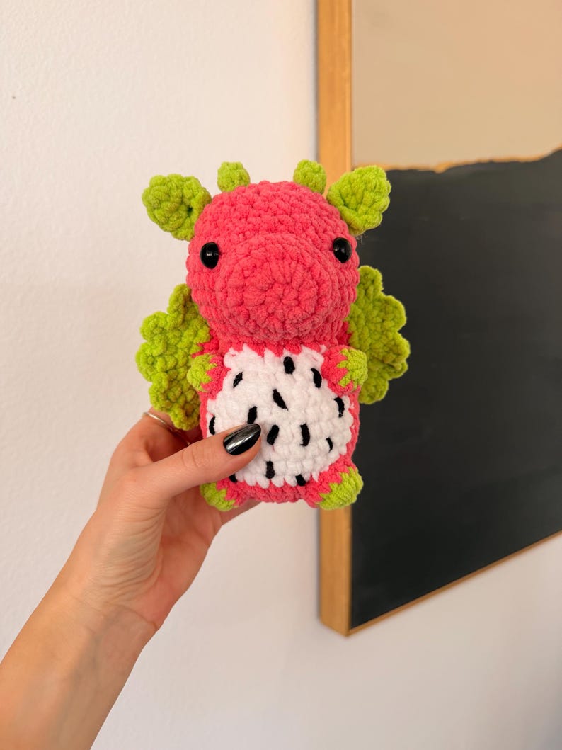 Puede incluir: Un peluche de drag&oacute;n hecho a mano con ganchillo en forma de fruta del drag&oacute;n. El peluche presenta un cuerpo rosa, alas y detalles verdes, y una barriga blanca con detalles de semillas negras. El drag&oacute;n tiene ojos de seguridad negros.
