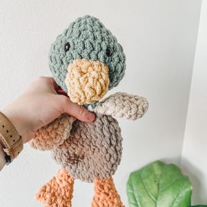 May include: A handmade crochet duck toy in shades of green, tan, brown, and orange. The duck has a textured body, a light green head, a tan beak, and orange feet. The toy is soft and plush, with black button eyes.
