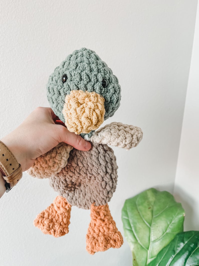 May include: A handmade crochet duck toy in shades of green, tan, brown, and orange. The duck has a textured body, a light green head, a tan beak, and orange feet. The toy is soft and plush, with black button eyes.