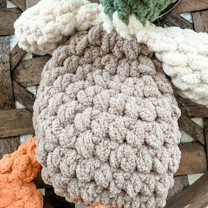May include: A handmade, plush crochet duck toy. The duck features a light brown body, white wings, and a green and yellow head. The feet are a soft orange color. The toy is made with a chunky yarn, giving it a textured appearance.