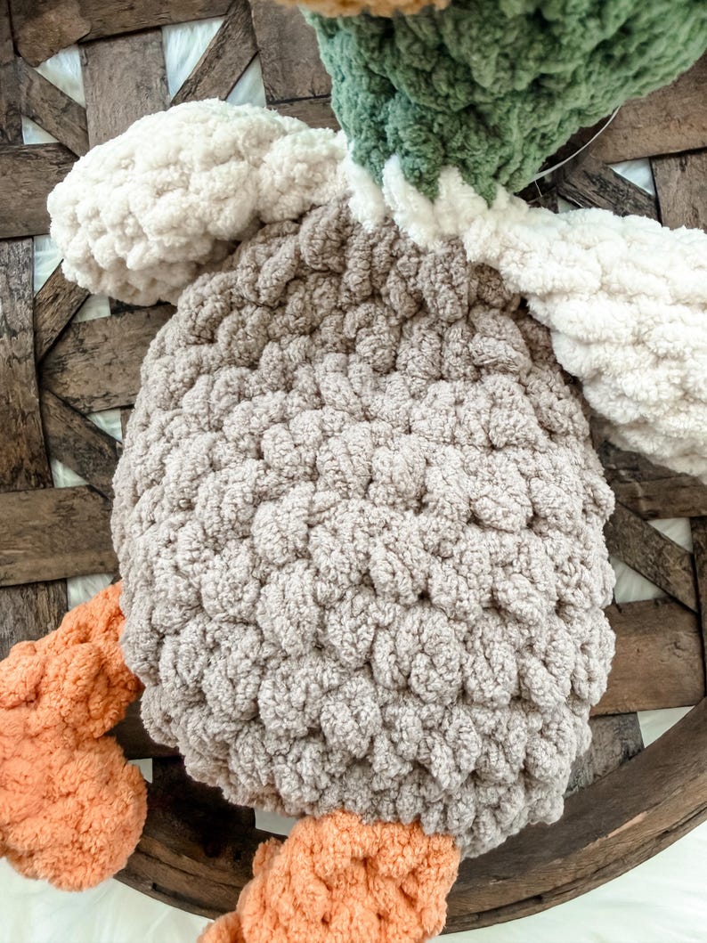 May include: A handmade, plush crochet duck toy. The duck features a light brown body, white wings, and a green and yellow head. The feet are a soft orange color. The toy is made with a chunky yarn, giving it a textured appearance.