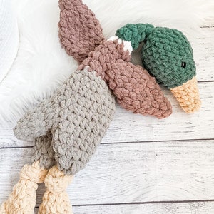 May include: A crocheted plush duck toy with a brown, gray, and green color scheme. The duck has a long, thin neck and a small, round head. The duck's feet are curled up and the duck is lying on its back.