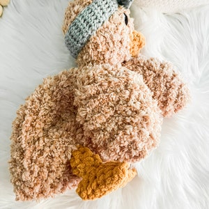 May include: A crocheted plush toy shaped like a duck. The duck is brown with a light blue crocheted headband. The duck is lying on a white fluffy surface.