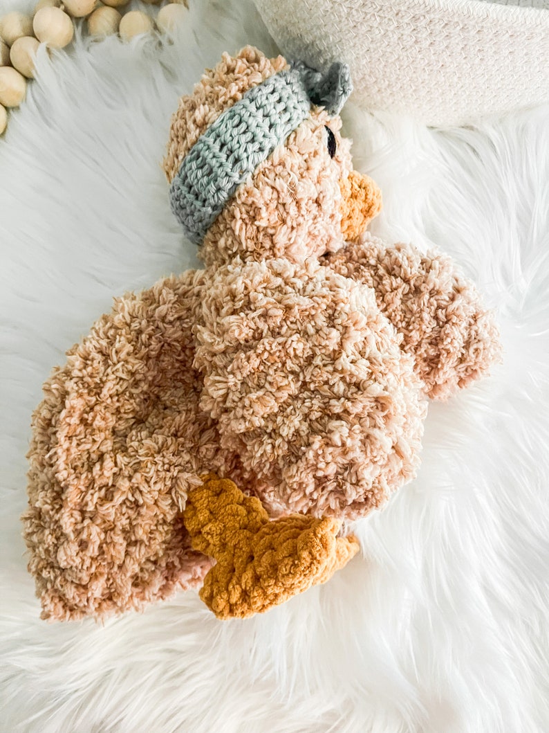 May include: A crocheted plush toy shaped like a duck. The duck is brown with a light blue crocheted headband. The duck is lying on a white fluffy surface.