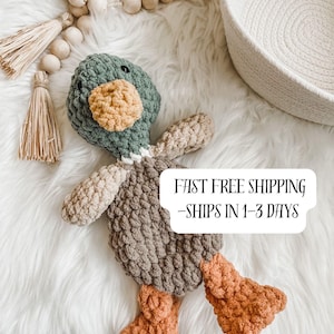 May include: A handmade crochet duck toy in shades of green, tan, brown, and orange. The duck has a green head, tan wings, a brown body, and orange feet. The toy is lying on a white, fluffy surface.