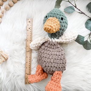 May include: A handmade crochet duck toy with a green head, orange beak and feet, and a gray body. The duck is approximately 10 inches tall, with white wings and is lying on a white fluffy surface. A wooden ruler is next to the duck.
