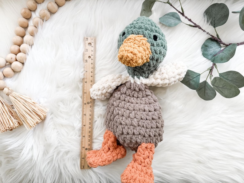 May include: A handmade crochet duck toy with a green head, orange beak and feet, and a gray body. The duck is approximately 10 inches tall, with white wings and is lying on a white fluffy surface. A wooden ruler is next to the duck.