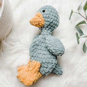 May include: A handmade, plush duck toy in shades of blue and yellow. The duck has a textured, soft body, with a light blue color. The beak and feet are a contrasting yellow-orange. The toy is designed to be a cuddly companion.