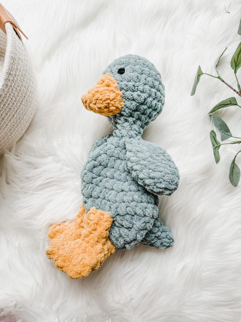May include: A handmade, plush duck toy in shades of blue and yellow. The duck has a textured, soft body, with a light blue color. The beak and feet are a contrasting yellow-orange. The toy is designed to be a cuddly companion.