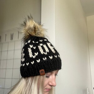 May include: A black knit beanie with a white geometric pattern spelling out the word "COVE". The hat features a large, fluffy, brown and tan faux fur pom-pom on top and a small brown leather tag.