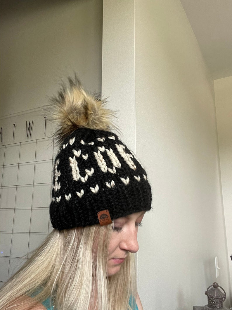 May include: A black knit beanie with a white geometric pattern spelling out the word "COVE". The hat features a large, fluffy, brown and tan faux fur pom-pom on top and a small brown leather tag.