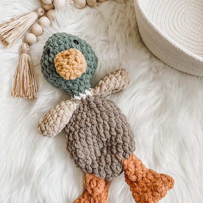 May include: A handmade crochet duck toy in shades of green, brown, tan, and orange. The duck has a textured body, a yellow beak, and orange feet. The toy is lying on a white, fluffy surface.