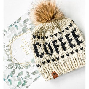 May include: Cream-colored knit beanie with the word "COFFEE" in black letters and a brown faux fur pom. The hat has a small brown leather tag. A "THANK YOU" card with a floral design is in the background.
