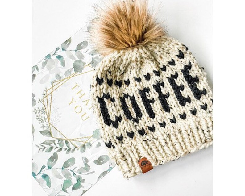 May include: Cream-colored knit beanie with the word "COFFEE" in black letters and a brown faux fur pom. The hat has a small brown leather tag. A "THANK YOU" card with a floral design is in the background.