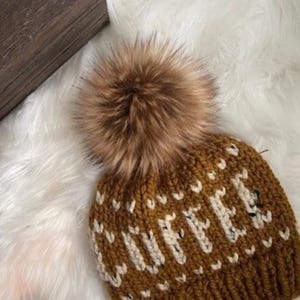 May include: A brown knit beanie with the word "COFFEE" in white letters. The hat has a large brown faux fur pom-pom on top. The beanie is displayed on a white, fluffy surface.