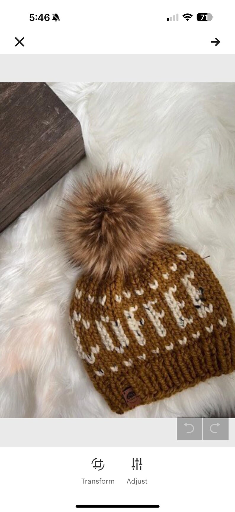 May include: A brown knit beanie with the word "COFFEE" in white letters. The hat has a large brown faux fur pom-pom on top. The beanie is displayed on a white, fluffy surface.