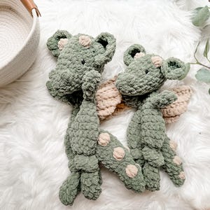 Peut inclure: Deux peluches dragons en crochet vert faites &agrave; la main, avec des ailes beiges et des taches rose clair. Les jouets doux sont pr&eacute;sent&eacute;s sur une surface blanche et moelleuse. Les dragons ont de petits yeux noirs et sont con&ccedil;us pour les enfants.