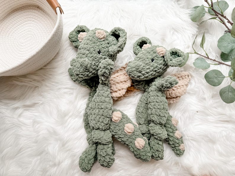 Peut inclure: Deux peluches dragons en crochet vert faites &agrave; la main, avec des ailes beiges et des taches rose clair. Les jouets doux sont pr&eacute;sent&eacute;s sur une surface blanche et moelleuse. Les dragons ont de petits yeux noirs et sont con&ccedil;us pour les enfants.