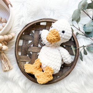 May include: A handmade white crochet duck with a yellow beak and feet sits in a rustic wooden basket. The duck has black eyes and a fluffy texture. The basket is on a white, fluffy surface.