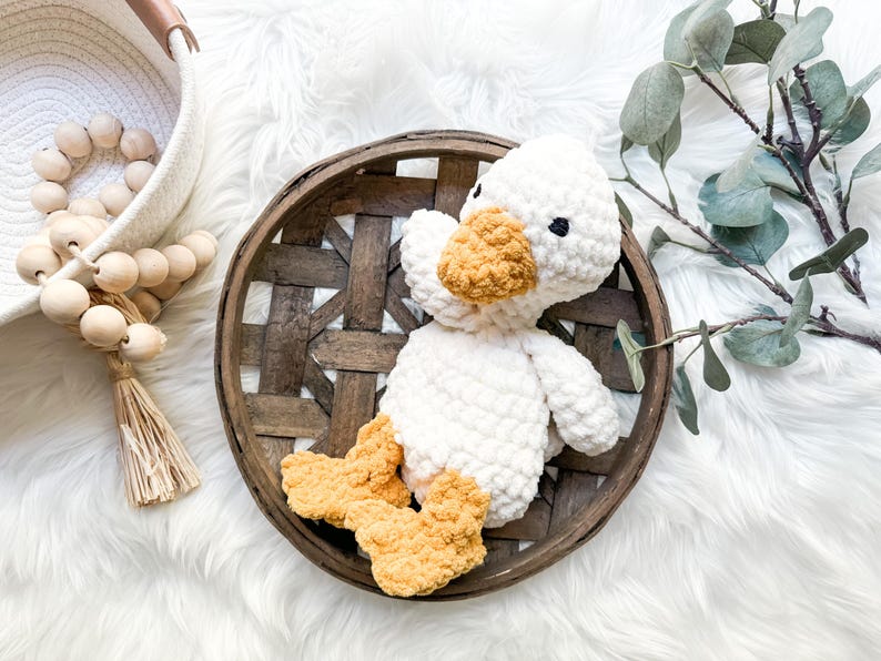 May include: A handmade white crochet duck with a yellow beak and feet sits in a rustic wooden basket. The duck has black eyes and a fluffy texture. The basket is on a white, fluffy surface.