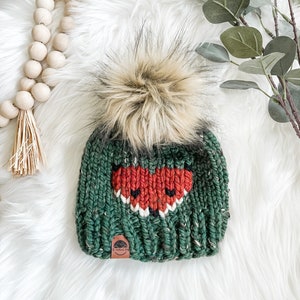May include: A green knit beanie with a fox face design in red, white, and black. The hat features a large, fluffy, brown faux fur pom-pom on top. A small, brown leather tag is sewn on the front.