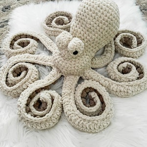 Pode incluir: Um brinquedo de polvo de croch&ecirc; com um corpo de cor creme e detalhes marrons. O polvo tem oito tent&aacute;culos que s&atilde;o enrolados em espiral.