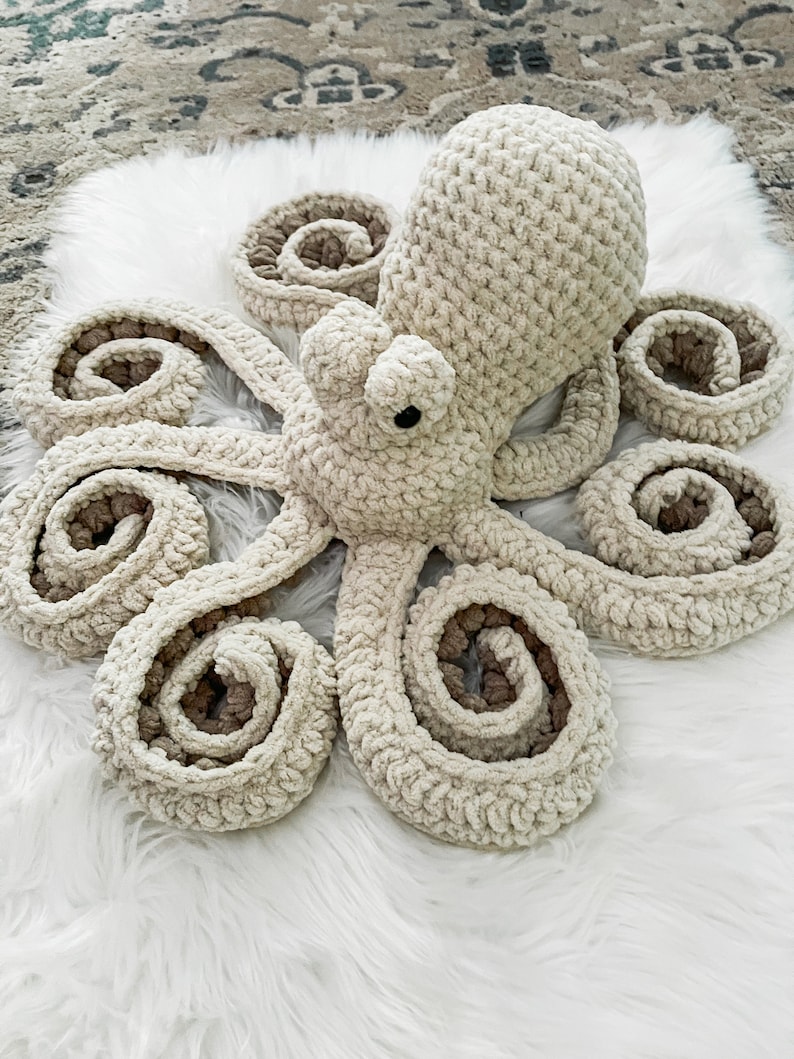 Pode incluir: Um brinquedo de polvo de croch&ecirc; com um corpo de cor creme e detalhes marrons. O polvo tem oito tent&aacute;culos que s&atilde;o enrolados em espiral.